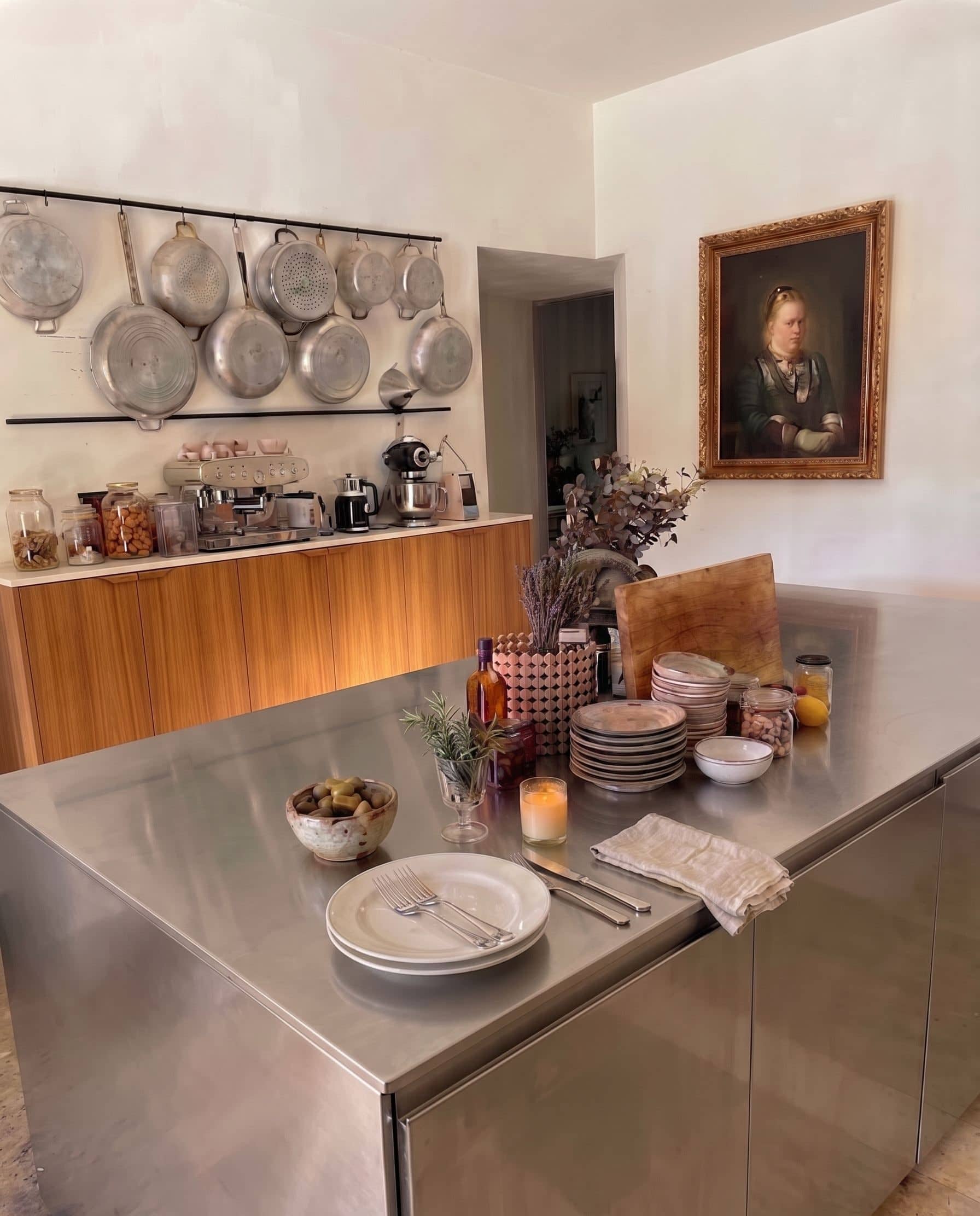 The Mamie kitchen - hanging aluminium pans, espresso machine, oil portrait of a matriarch on the wall, stainless steel island laid with plates, candle, and a small bowl.